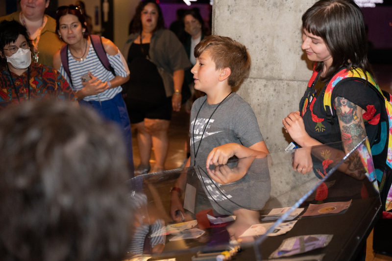 A young boy pointing at a display case and giving a presentation while a group of adults watch, during a MOPOP summer camp