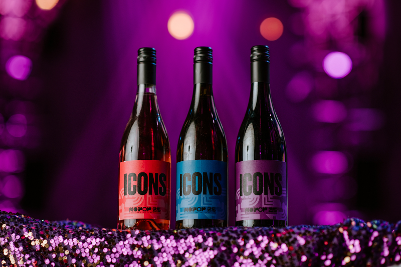Three wine bottles labeled ICONS with red, blue, and purple labels on a sequined surface against a purple blurred background.