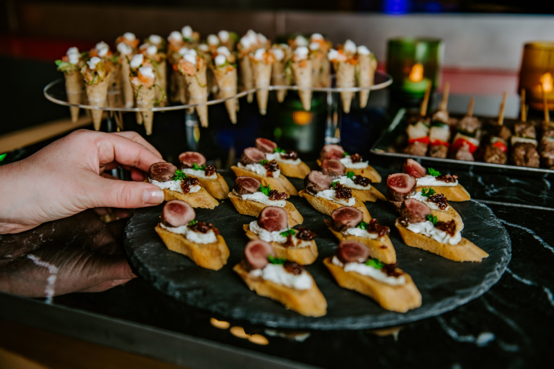 Hand picking a canapé topped with cream, meat slices, and garnish from a black plate with multiple similar canapés, with additional appetizers in the background.