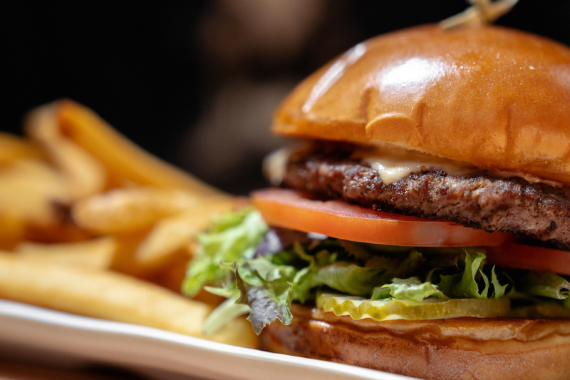 Close-up of a cheeseburger with lettuce, tomato, and pickles served with French fries on a white plate.
