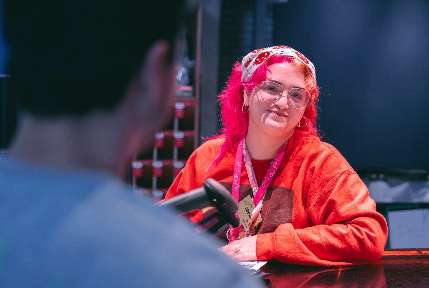 Smiling staff member with bright pink hair and glasses wearing red sweater and MoPOP lanyard behind a counter.