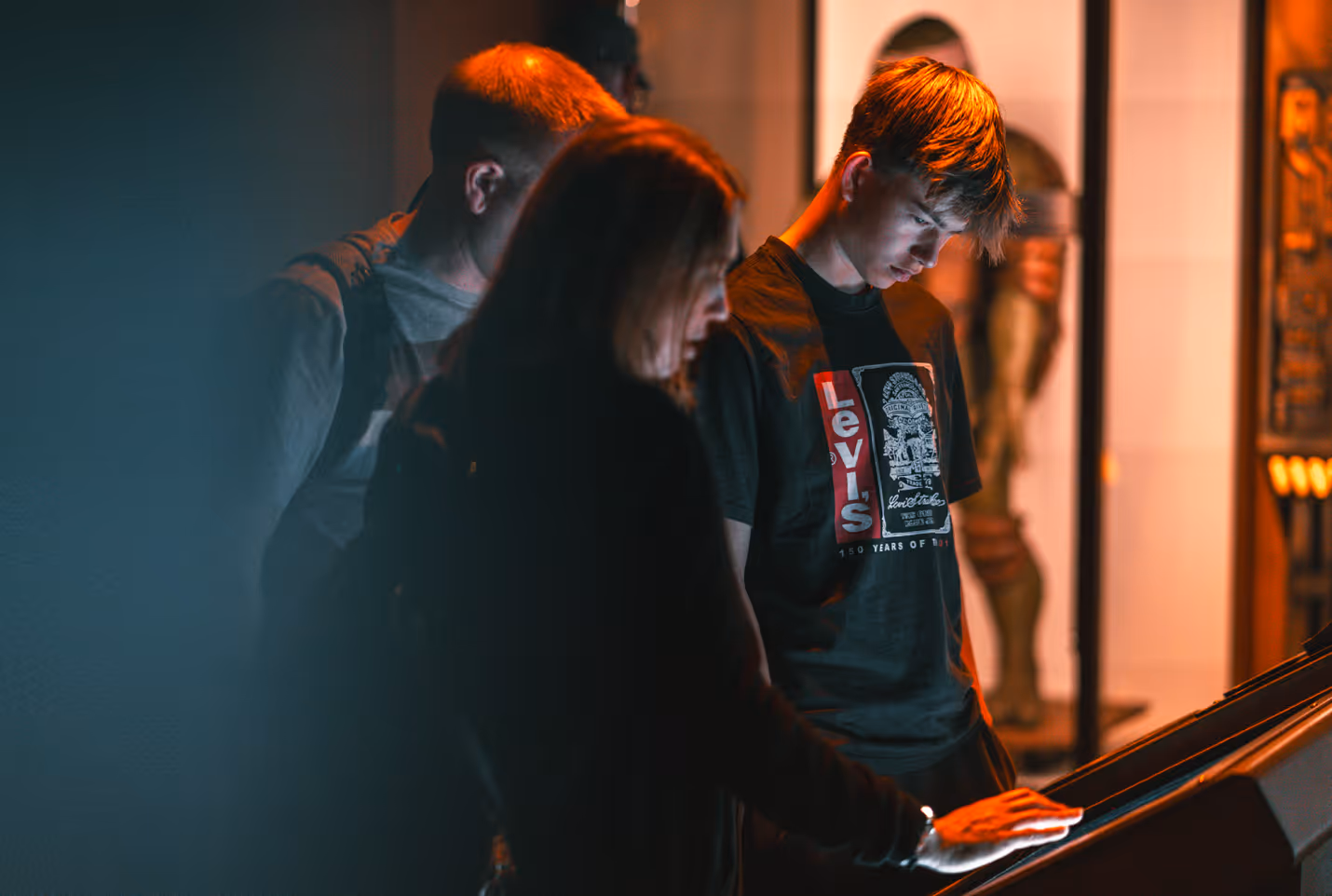 Three people closely looking at and interacting with a touchscreen display in a dimly lit museum setting.