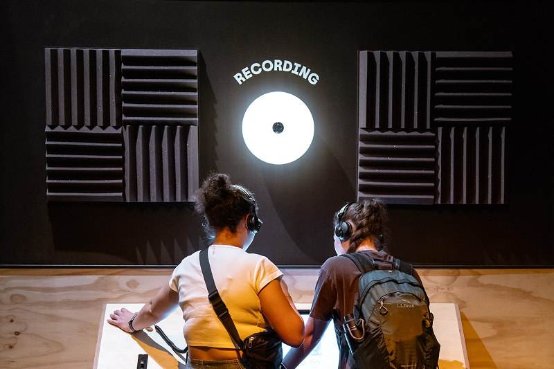 Two people wearing headphones interacting with a tabletop display under a lit sign that says 'RECORDING' with a vinyl record graphic on a black wall with acoustic foam panels.