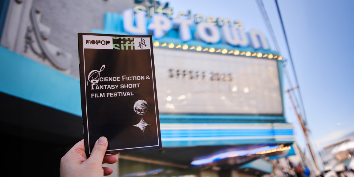 A hand holds a Science Fiction & Fantasy Short Film Festival program in front of a blurred Uptown cinema marquee.