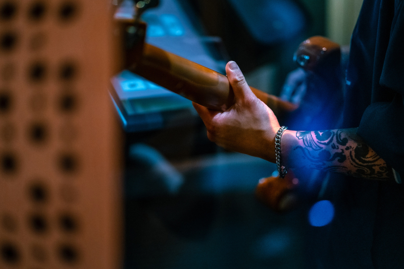 Close-up of a tattooed arm and hand playing a bass guitar in a dimly lit music studio.