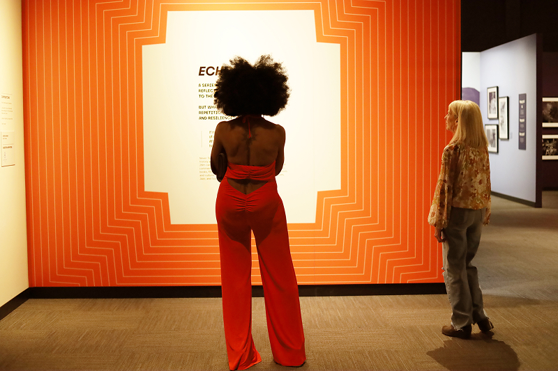A Black woman with an afro, wearing a red jumpsuit, and a white blonde woman read an illuminated sign in the Never Turn Back exhibition