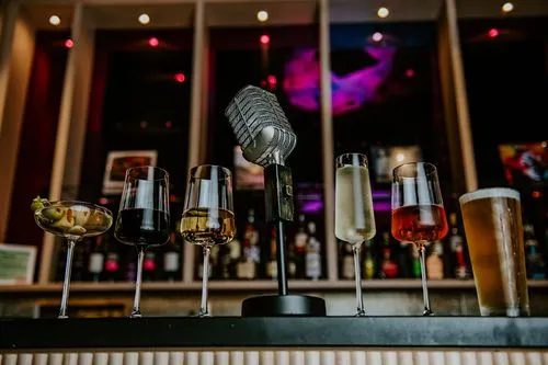 Vintage microphone surrounded by six different drinks on a bar counter with a blurred background.