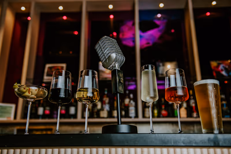 Vintage microphone surrounded by six different drinks on a bar counter with a blurred background.