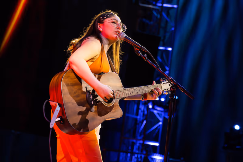Female musician wearing glasses and orange outfit playing acoustic guitar and singing on stage.