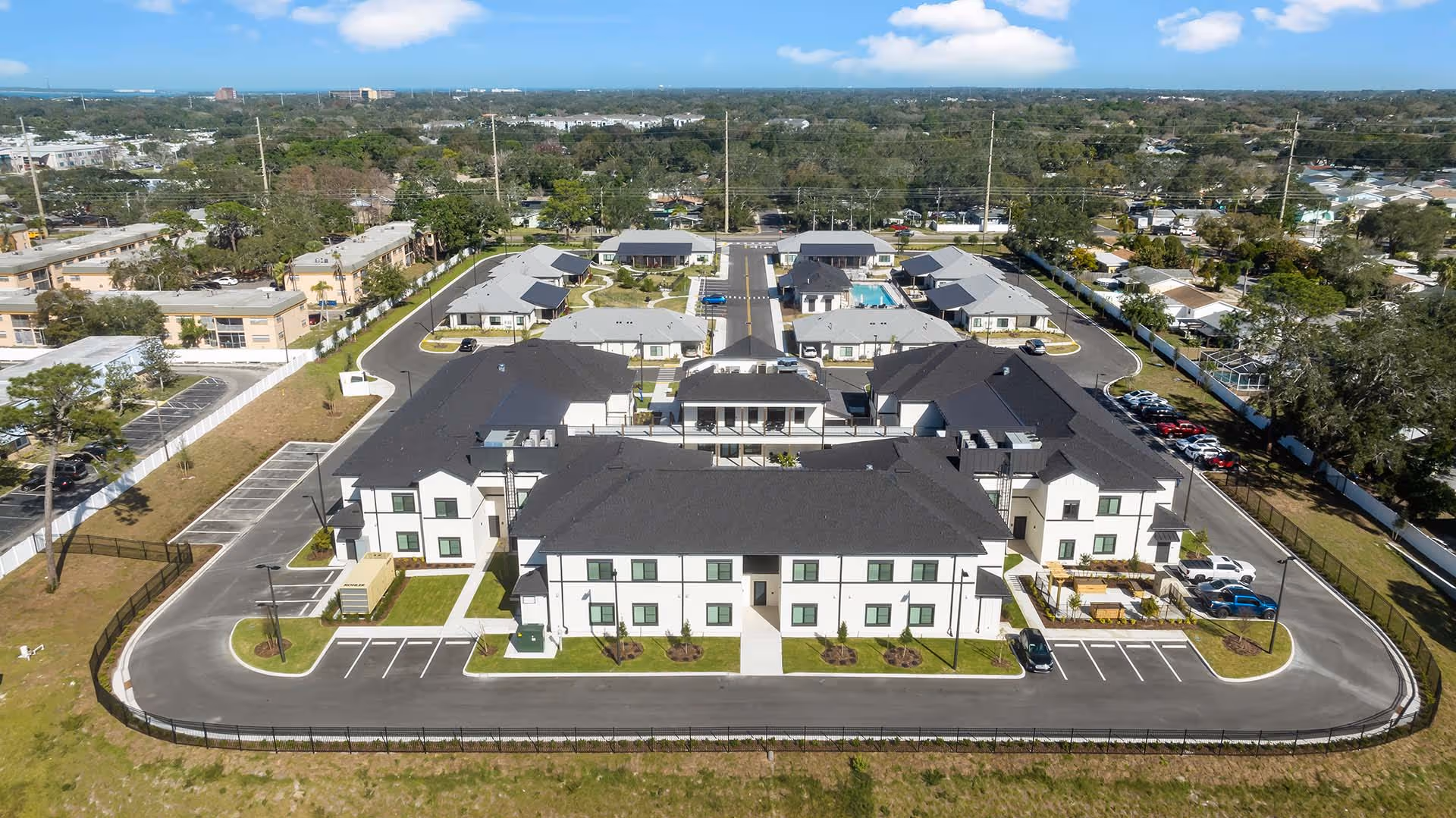 Aerial view of a modern residential apartment complex with black roofs, white walls, parking spaces, and surrounding greenery.