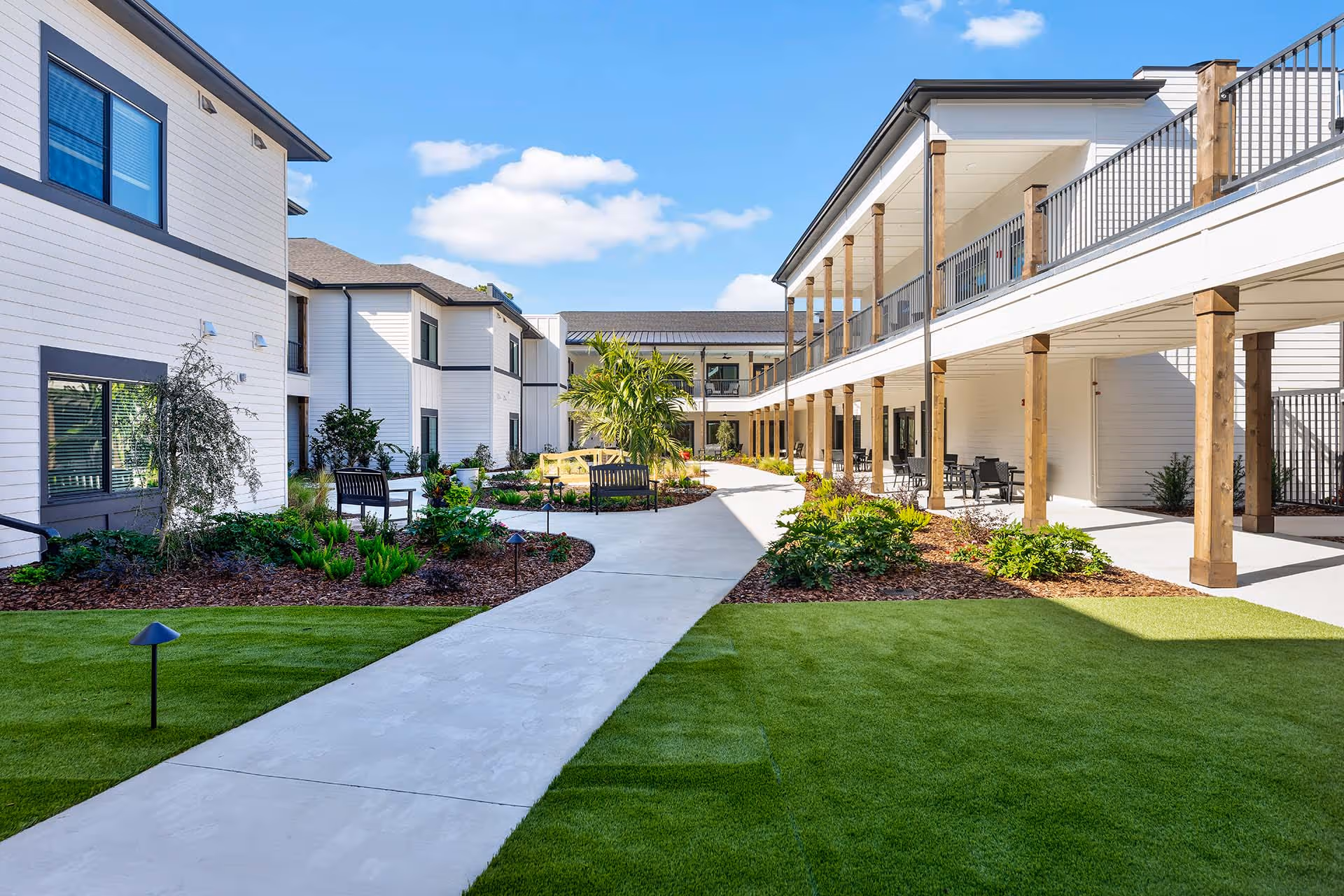 Modern apartment complex courtyard with landscaped gardens, benches, and a central concrete walkway under a bright blue sky.