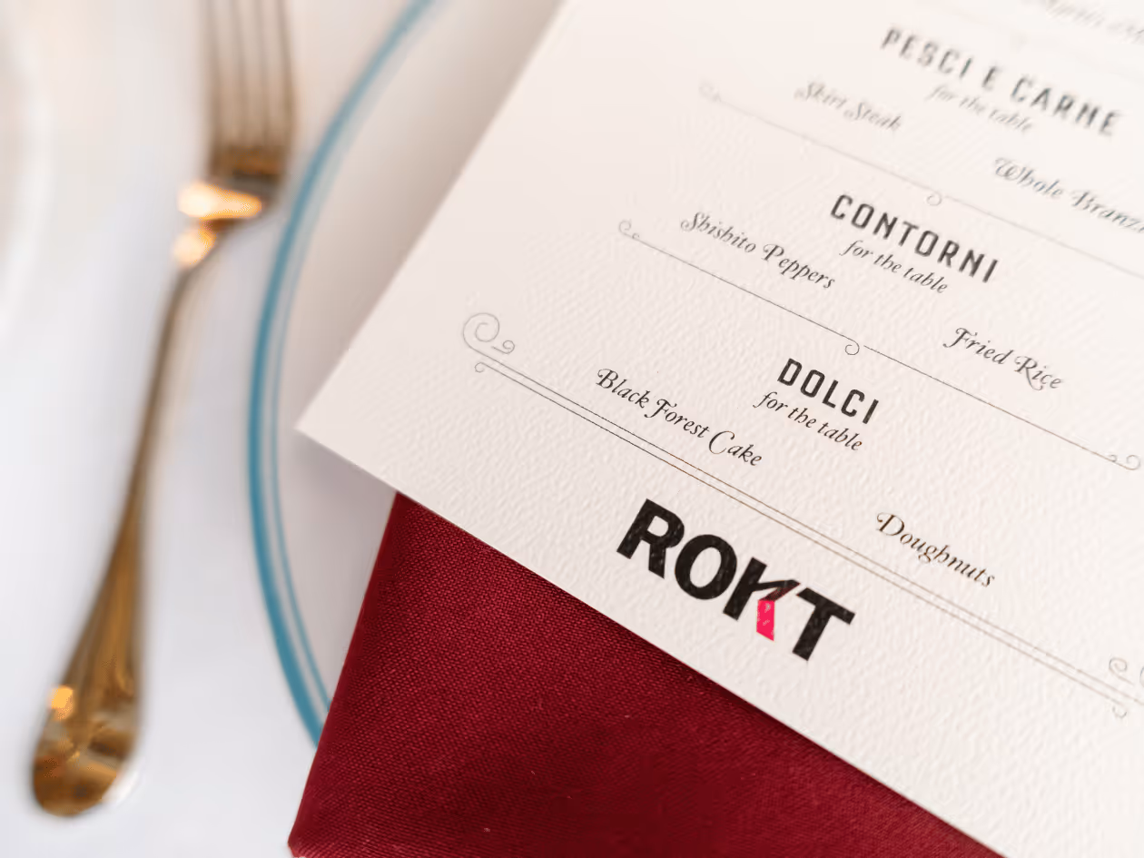 A close-up shot of an elegant printed dinner menu placed over a deep red cloth napkin on a white plate with a light blue rim. The menu, printed on textured white cardstock, lists shared items under "Contorni" (shishito peppers and fried rice) and "Dolci" (Black Forest cake and doughnuts). At the bottom, the logo "ROKT" is prominently displayed in bold black letters, with the letter "K" stylized in pink. A gold-toned fork is partially visible to the left, adding to the upscale table setting.