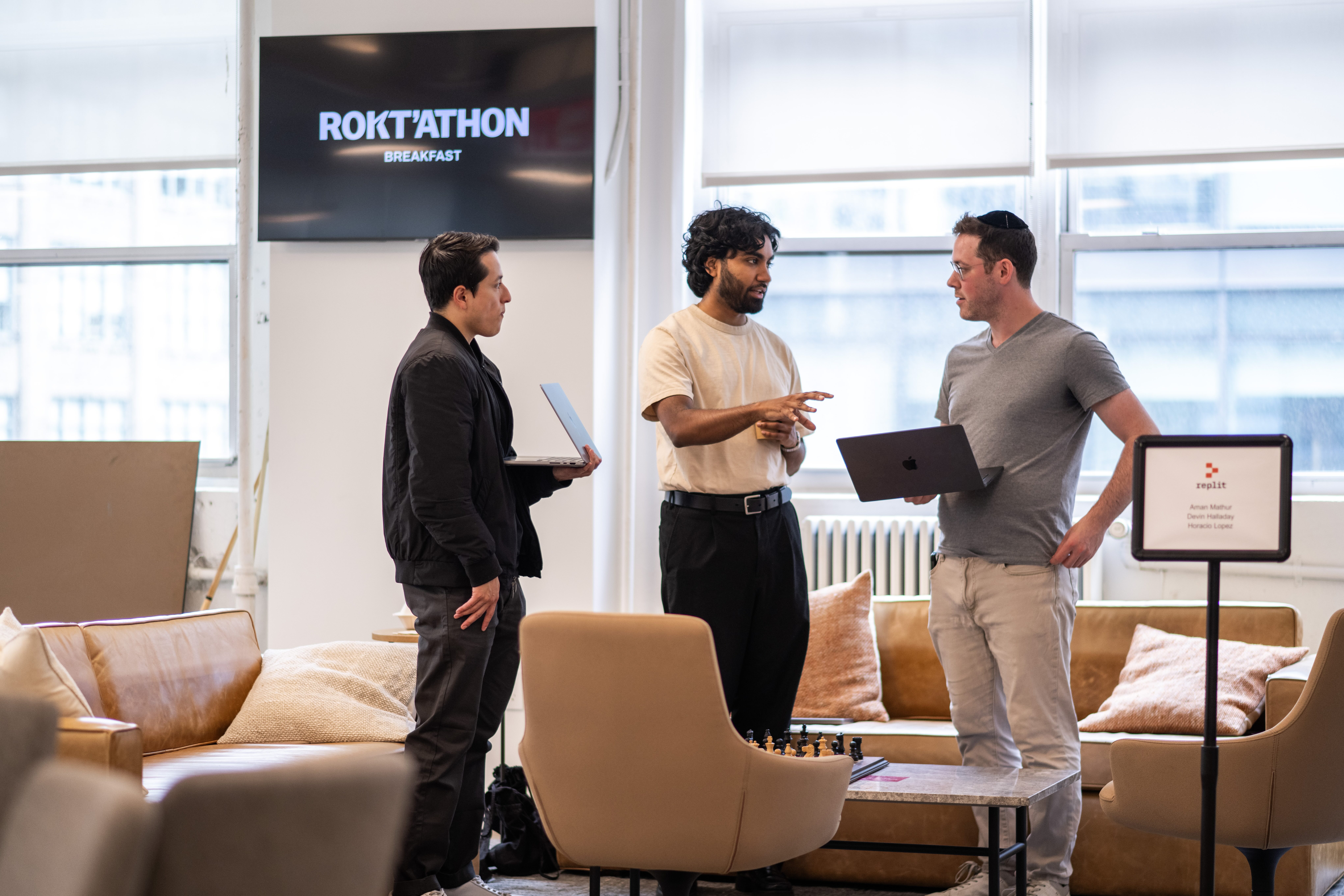 Three men stand in discussion during a tech event called “ROKT’ATHON,” each holding laptops in a casual lounge area with modern furniture and a chessboard on the table.
