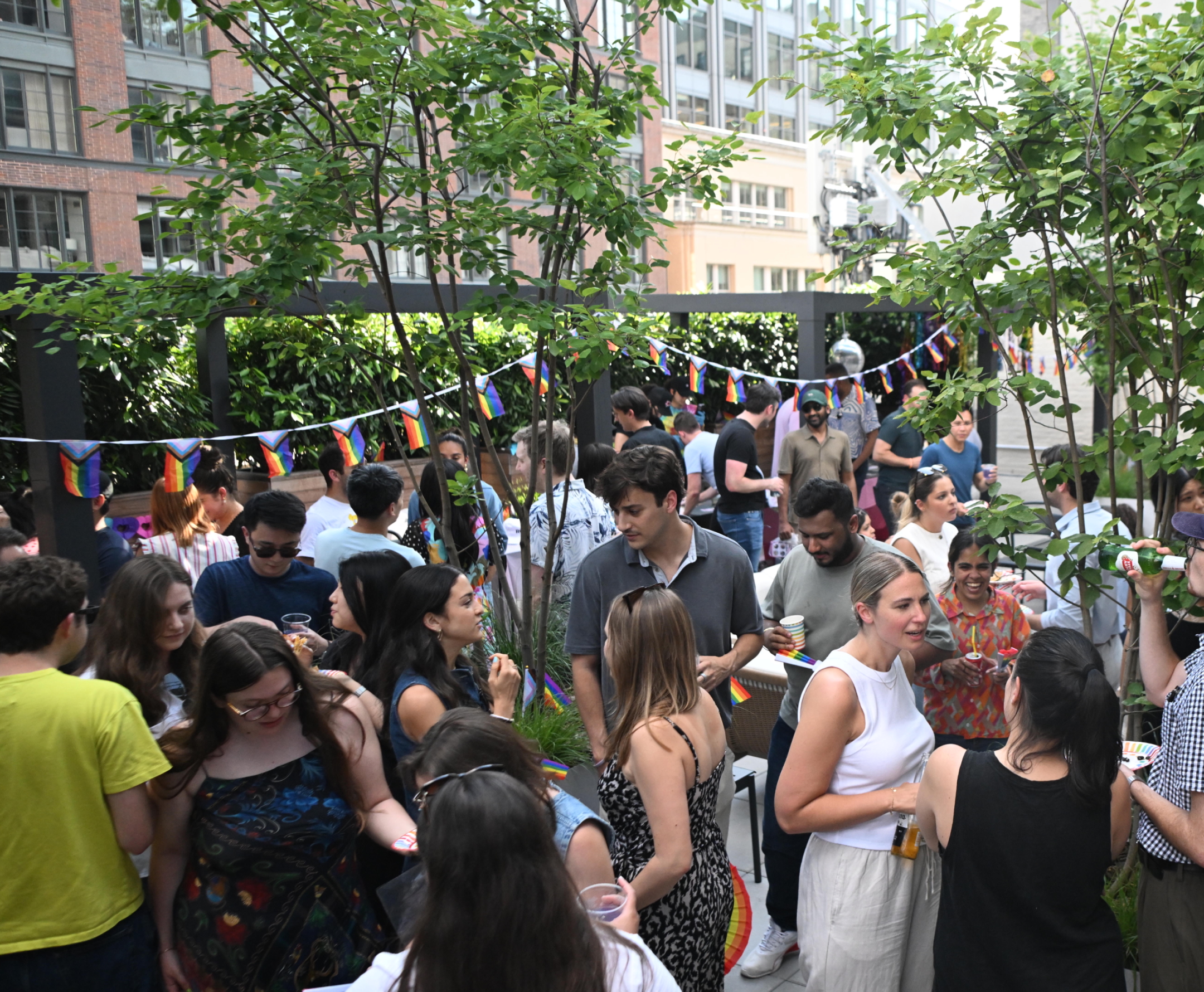 A lively rooftop party with people socializing among greenery, under strings of rainbow Pride flags. Guests are chatting, holding drinks, and enjoying a warm, festive outdoor atmosphere.