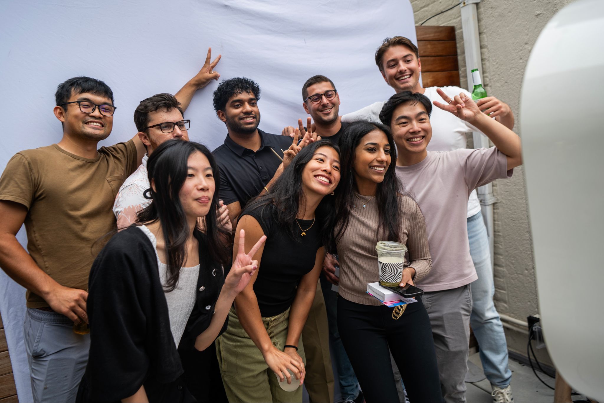 A cheerful group of young adults poses for a photo in front of a white backdrop, many flashing peace signs and smiling. The setting appears casual and social on the Rokt NYC Office Rooftop..