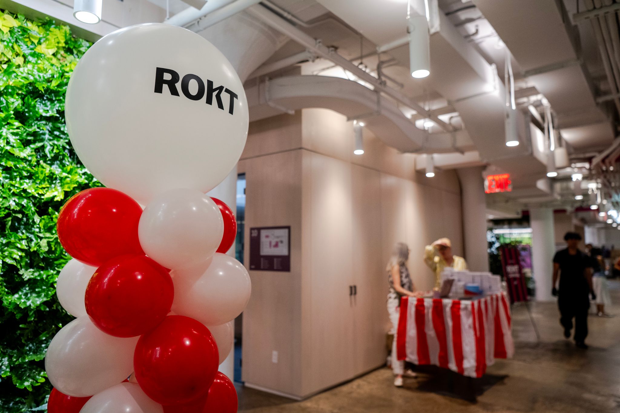 A large balloon display with the ROKT logo stands in a brightly lit office hallway, near a red-and-white striped table where two people are chatting.