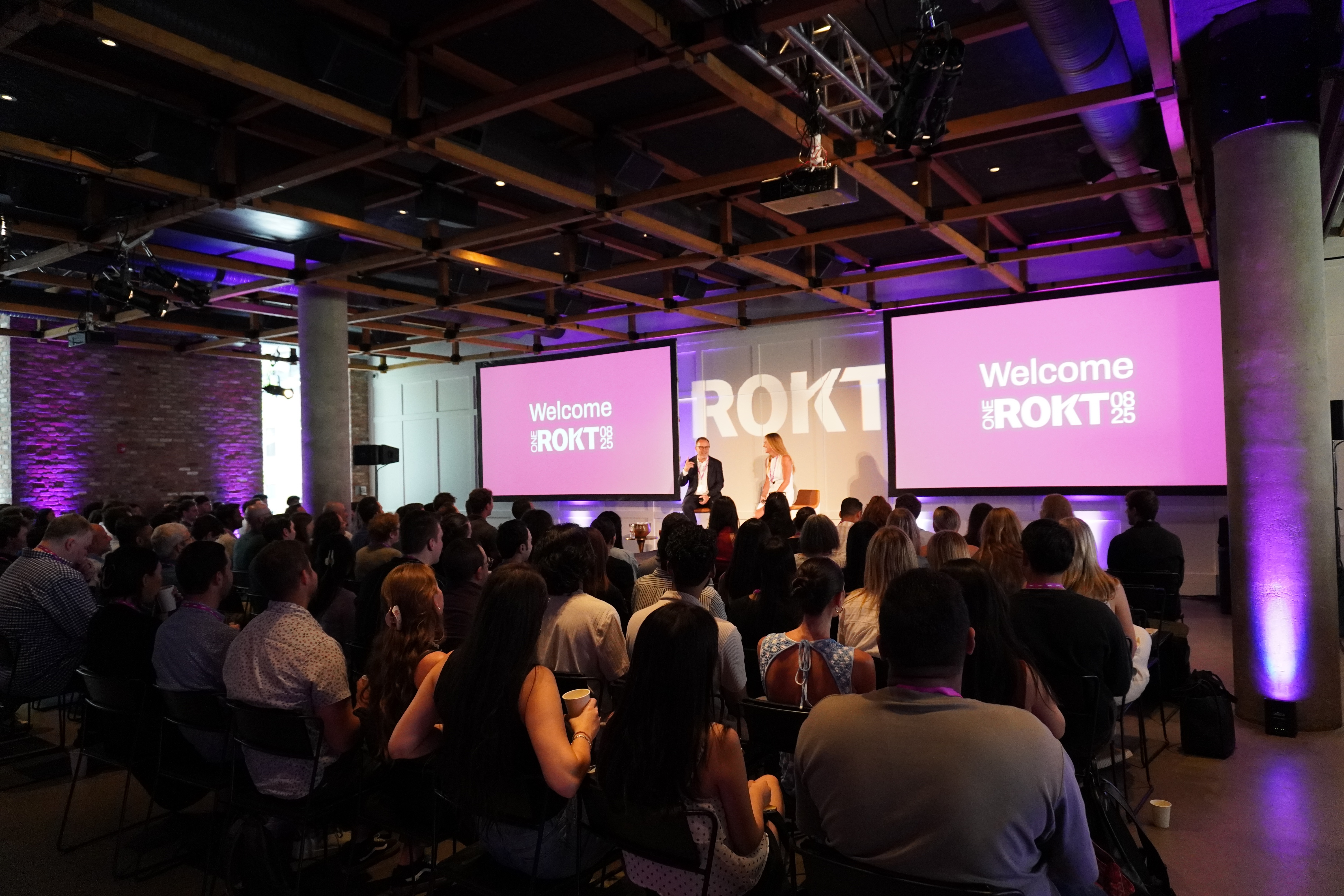 A large audience sits facing a stage with two speakers and dual screens displaying “Welcome ONE ROKT 08 25” in bold pink branding, at a corporate event in a modern, industrial-style venue.