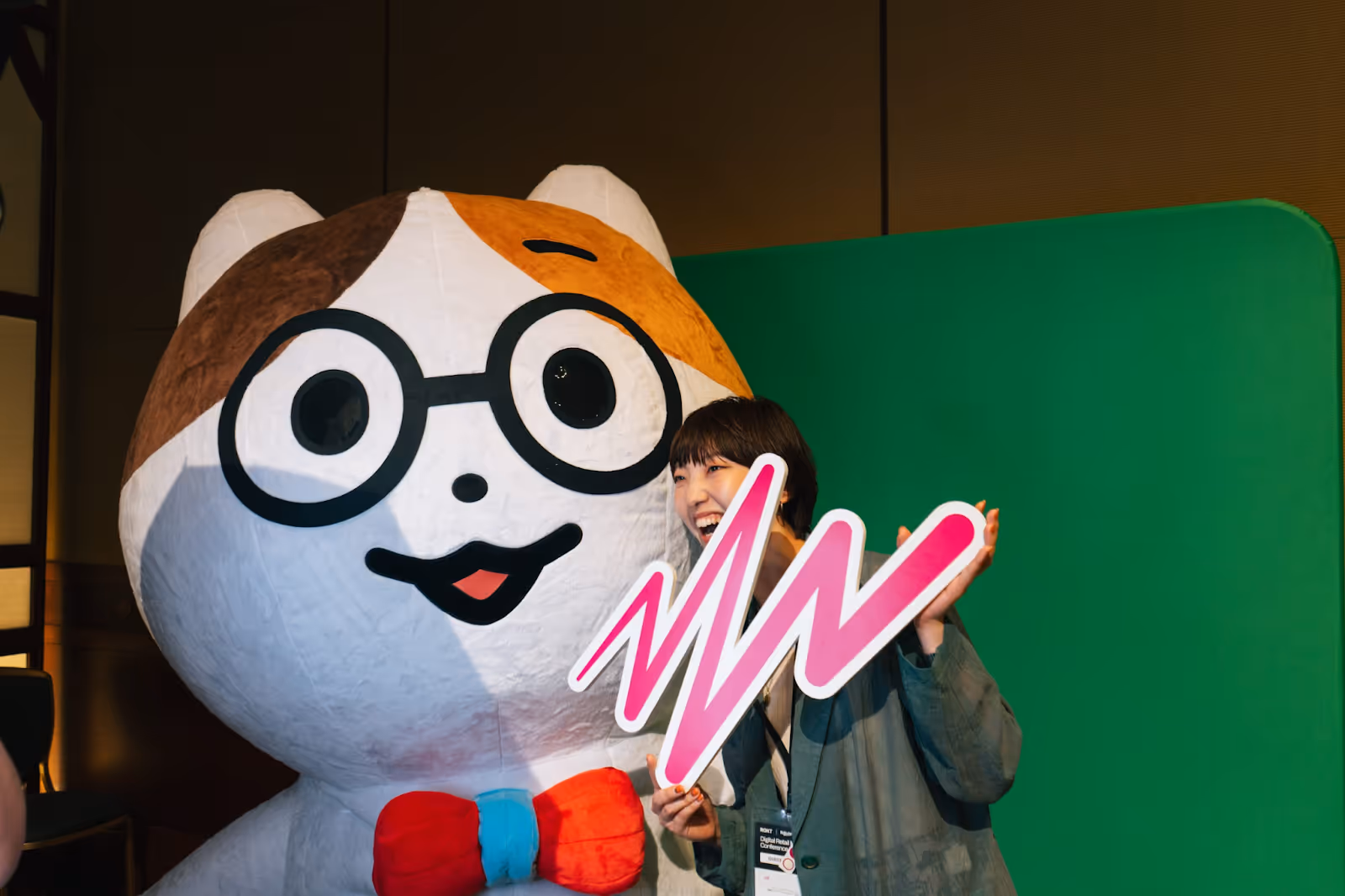 A cheerful woman poses next to a large, cartoonish cat mascot wearing oversized black glasses and a red-and-blue bow tie. The mascot has a white body with brown patches and a wide, smiling face. The woman is laughing and holding a bold, pink zigzag sign, partially covering her mouth. Behind them is a green backdrop from the photobooth at the event.