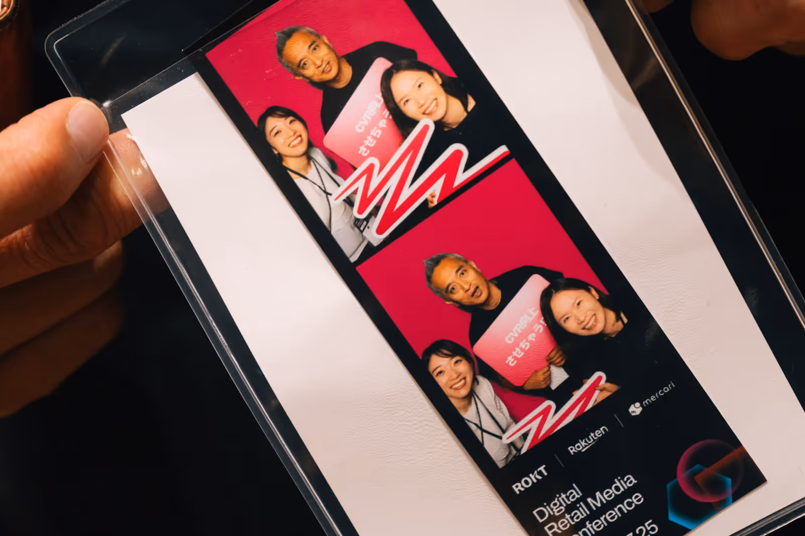 A close-up of a laminated photo booth strip featuring three smiling people posing in front of a bright pink backdrop. The group includes two women and a man holding a Rokt Connector prop. The man wears a shirt with text in English and Japanese. The bottom of the strip displays branding for ROKT, Rakuten, and Mercari, along with the event name: Digital Retail Media Conference 2025.