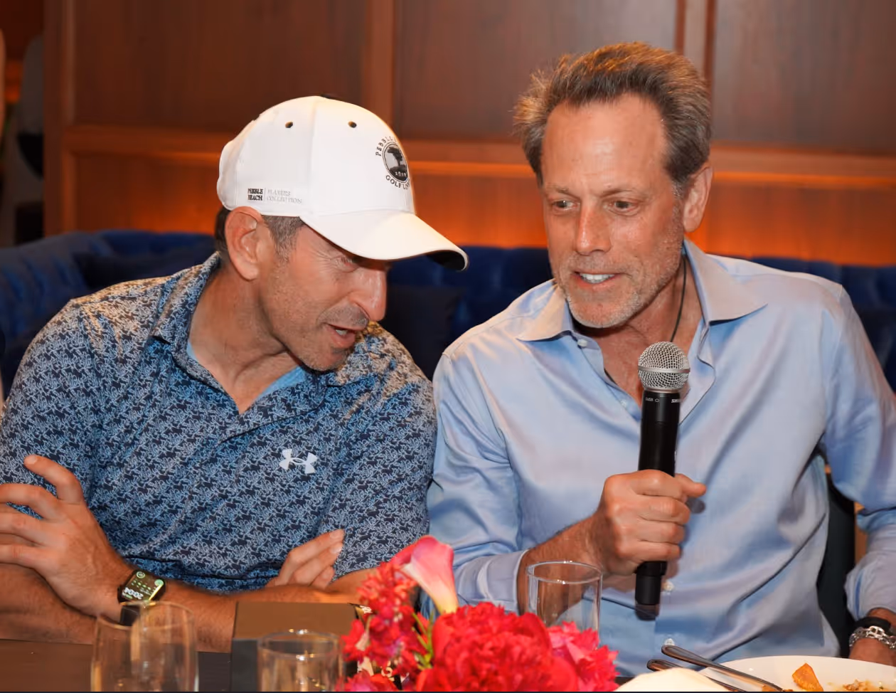 Two men sit closely at a dinner table, engaged in conversation. The man on the left wears a patterned blue Under Armour polo shirt and a white baseball cap with “Pebble Beach Golf Links” branding. The man on the right, in a light blue dress shirt, holds a microphone and appears to be speaking while leaning in. A bouquet of bright pink flowers are visible on the table in front of them while they sit at the dinner portion of the event.
