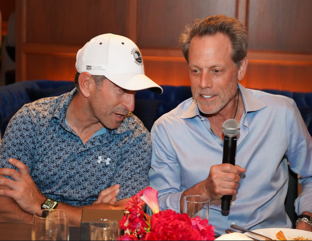 Two men sit closely at a dinner table, engaged in conversation. The man on the left wears a patterned blue Under Armour polo shirt and a white baseball cap with “Pebble Beach Golf Links” branding. The man on the right, in a light blue dress shirt, holds a microphone and appears to be speaking while leaning in. A bouquet of bright pink flowers are visible on the table in front of them while they sit at the dinner portion of the event.