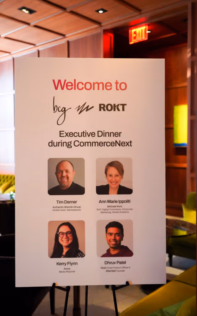 A standing event sign welcomes attendees to the “bcg x ROKT Executive Dinner during CommerceNext.” The poster features headshots and names of four speakers: Tim Denrer (Authentic Brands Group), Ann Marie Ippoliti (Michael Kors), Kerry Flynn (Axios), and Dhruv Patel (Rokt and Alter/Sell). The sign is set against the backdrop of a stylish lounge with wood-paneled walls, soft lighting, and green velvet seating.