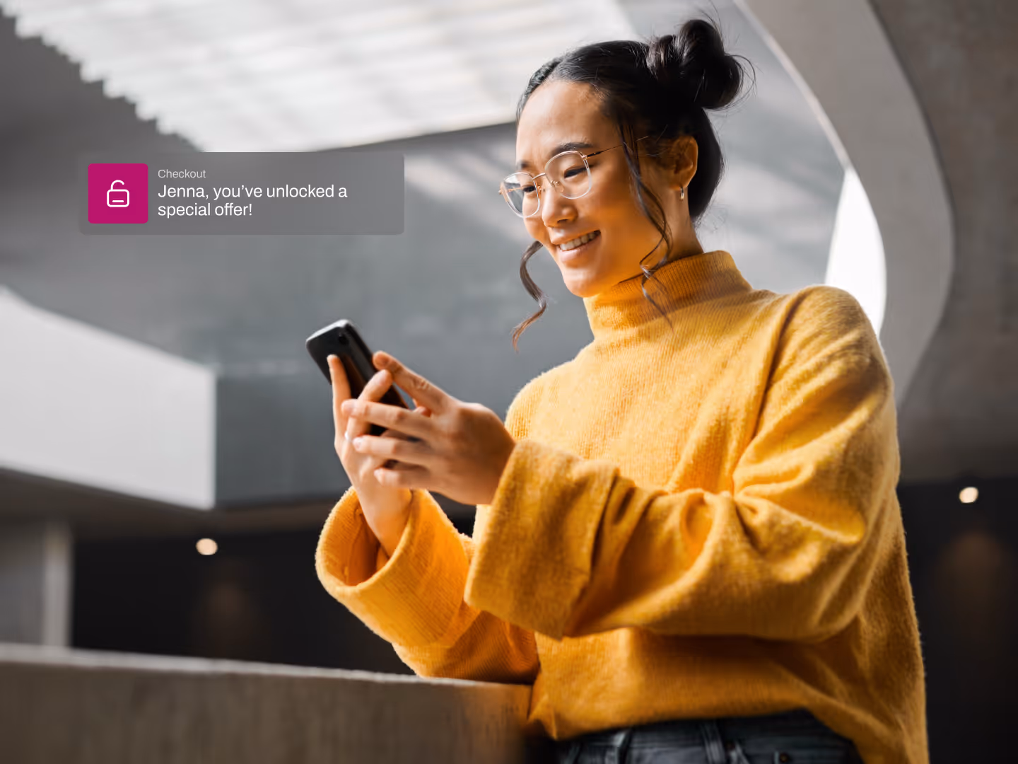 A young woman in a yellow turtleneck smiles at her phone in a modern building, her hair in two buns and round glasses framing her face.