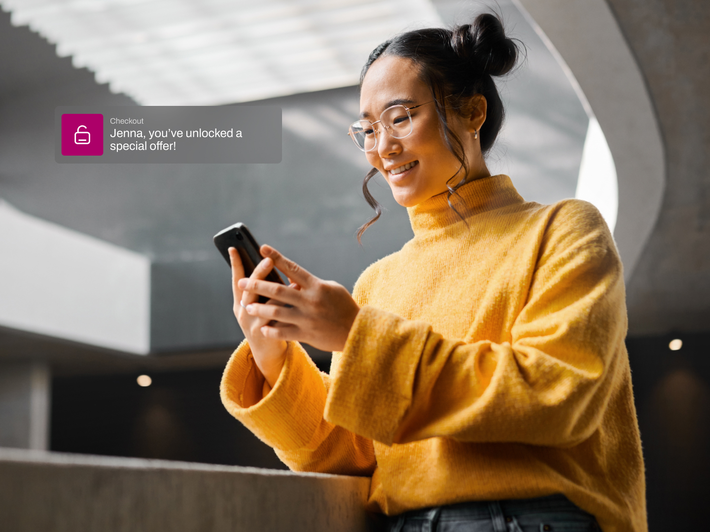A young woman in a yellow turtleneck smiles at her phone in a modern building, her hair in two buns and round glasses framing her face.