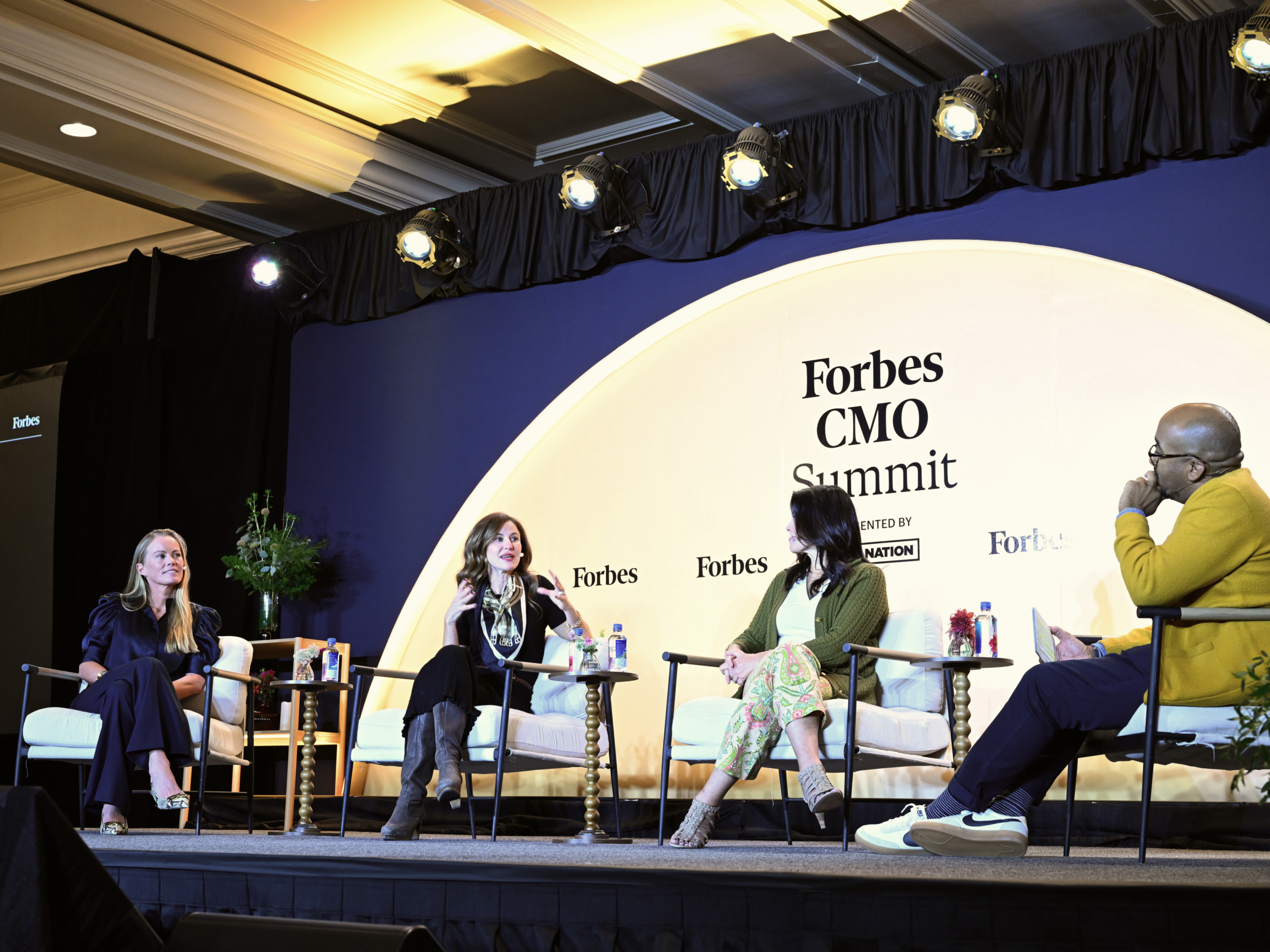 Four speakers sit on stage at the Forbes CMO Summit, engaged in a panel discussion on marketing strategy. The setting features a branded backdrop, modern chairs, and small tables with water and flowers.