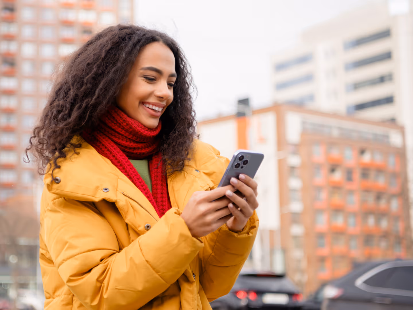 A young woman in a bright yellow puffer jacket and red knit scarf smiles while looking at her smartphone. She stands outdoors in a city setting with tall buildings in the background, suggesting a chilly day. Her body language and expression convey joy and 