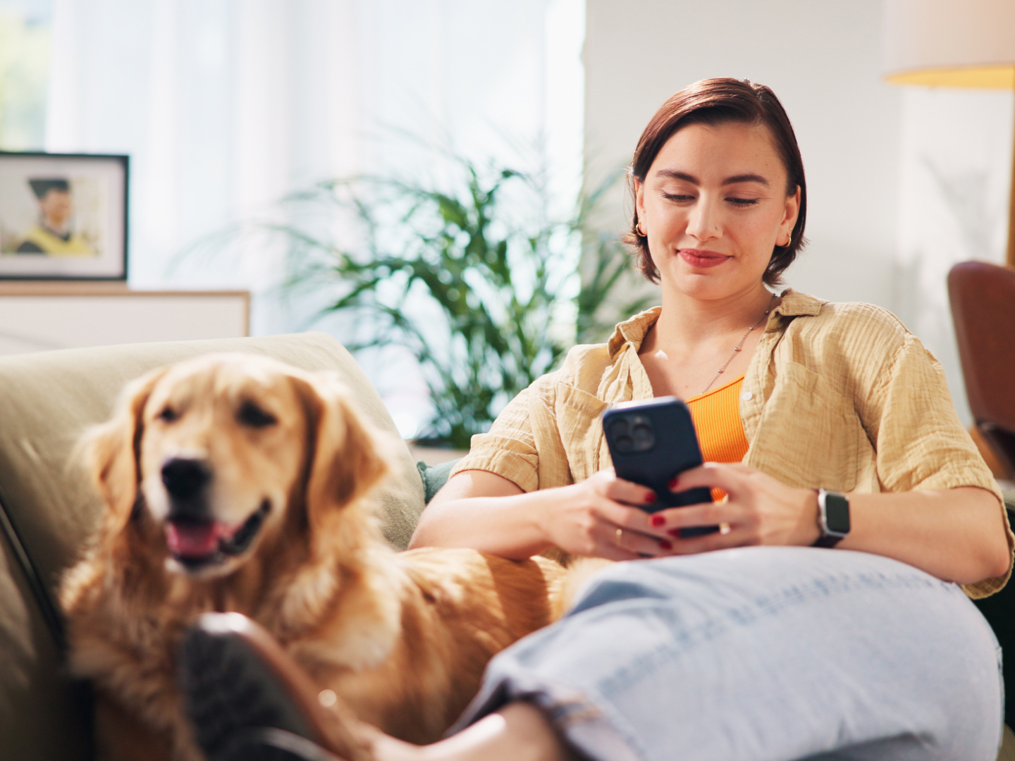 A woman relaxes on a couch at home, smiling as she looks at her smartphone. A golden retriever sits beside her with its tongue out, adding to the cozy, content atmosphere. Sunlight filters through a window behind them, highlighting a casual, peaceful momen
