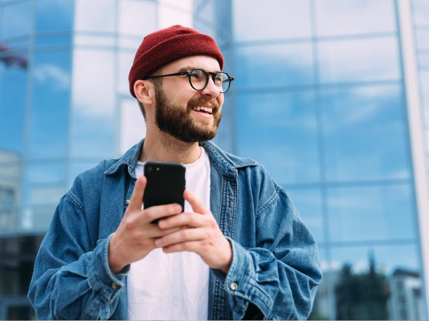 A smiling man in a red beanie and denim jacket holds a smartphone outdoors in front of a glass office building.