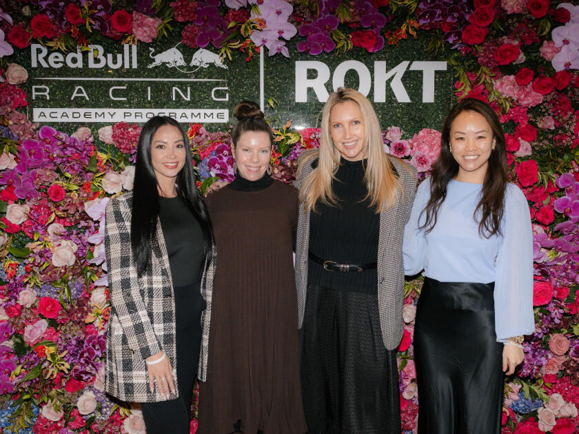 Four women pose together smiling in front of a floral wall backdrop featuring the Red Bull Racing Academy Programme and Rokt logos.