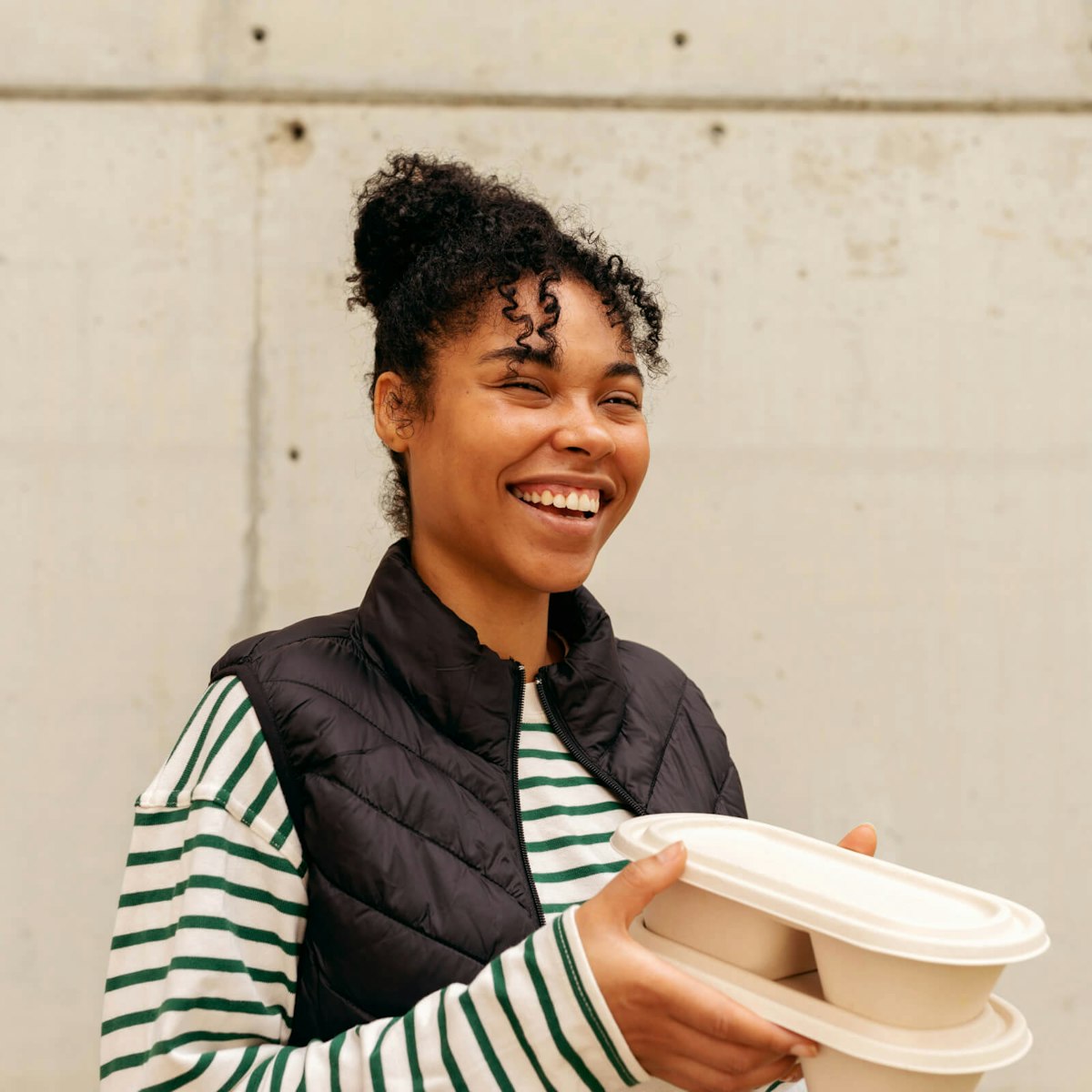 A smiling woman with curly hair tied up, wearing a black quilted vest over a green-and-white striped shirt, holds a beige compostable food container. She stands in front of a light-colored concrete wall.