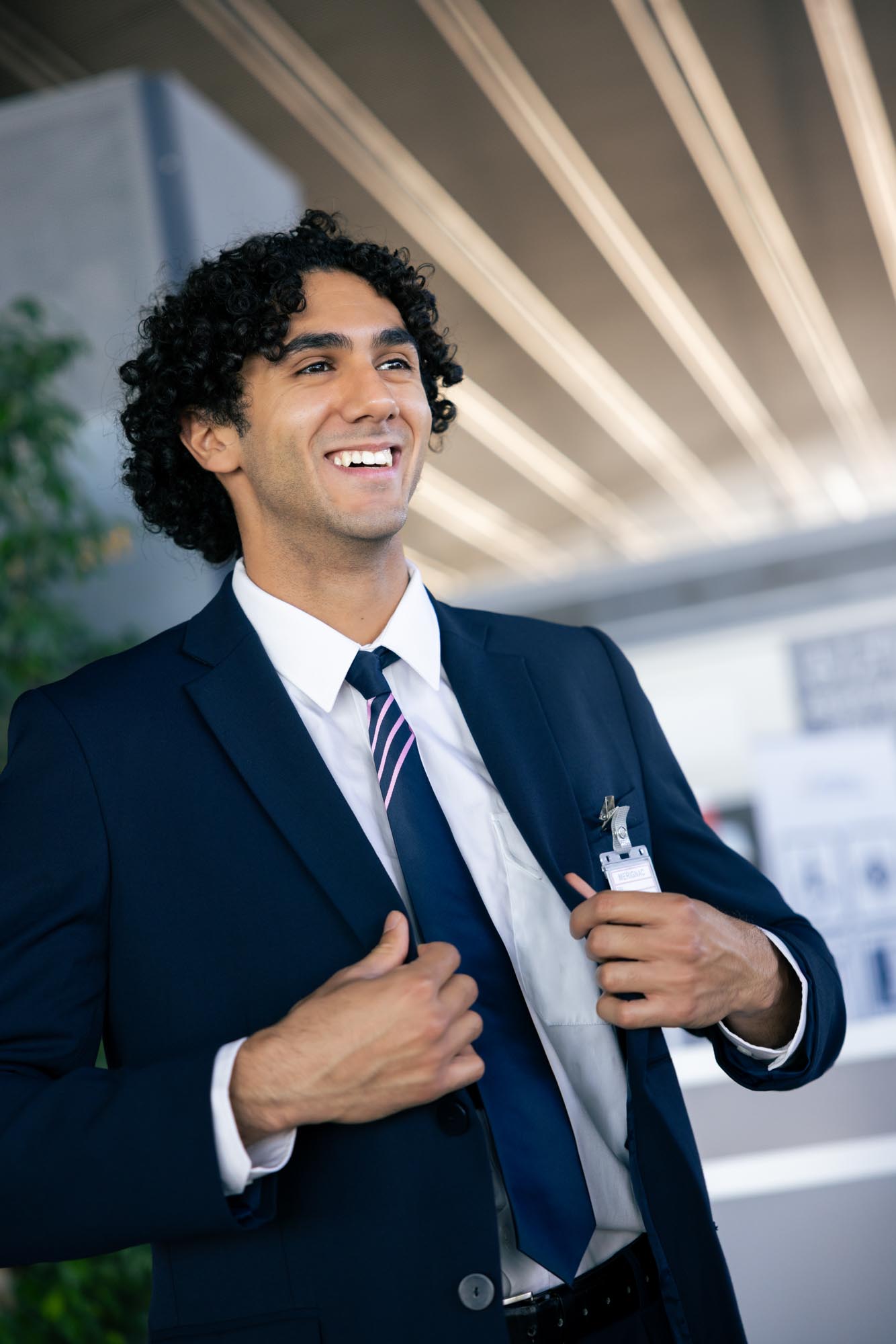 Jeune homme souriant en costume bleu foncé ajustant sa veste dans un environnement professionnel moderne.