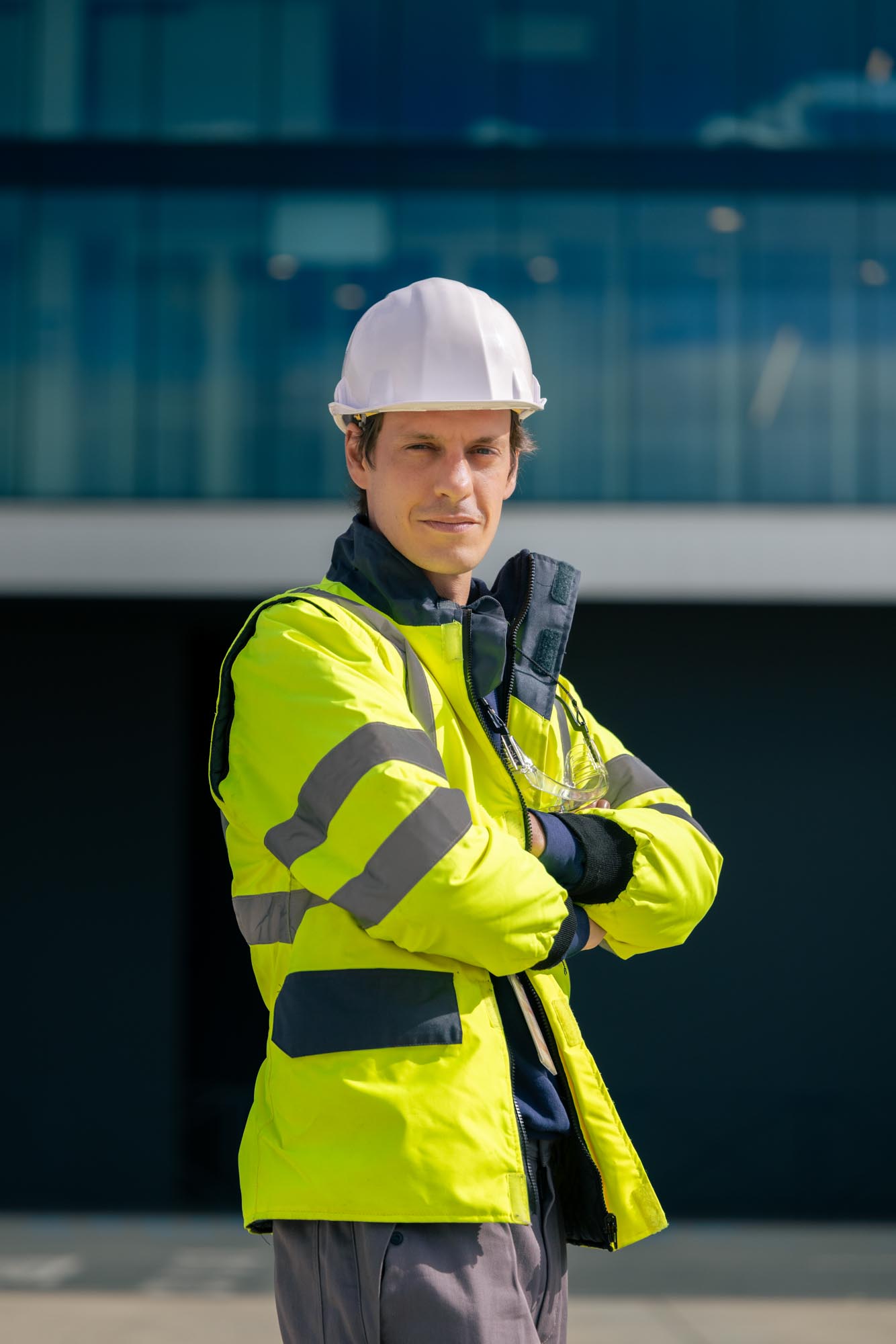 Homme portant un casque blanc et une veste de haute visibilité jaune avec bandes réfléchissantes, bras croisés devant un bâtiment moderne.