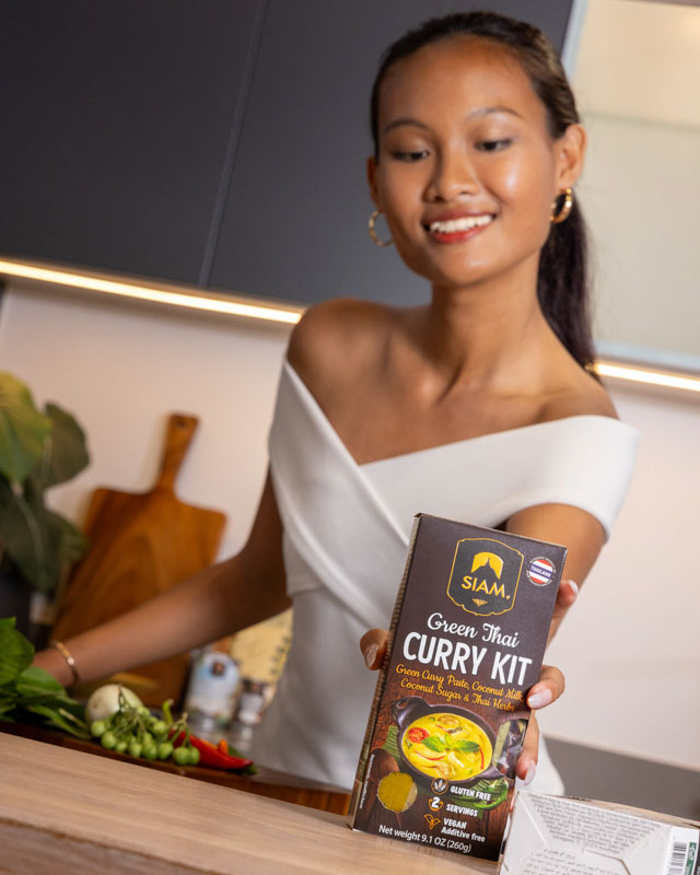 Une femme souriante tenant un kit de curry thaï vert de la marque SIAM dans une cuisine moderne.