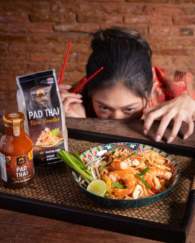 Une femme avec des baguettes rouges regarde un bol de Pad Thai aux crevettes, accompagné d'une bouteille de sauce et d'un paquet de nouilles de riz Pad Thai sur un plateau noir.