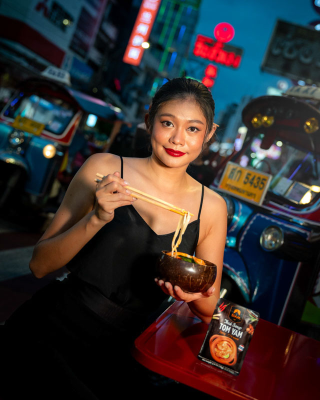 Jeune femme asiatique mangeant des nouilles dans un bol en bois avec des baguettes, assise dans une rue animée de nuit à Bangkok, avec un tuk-tuk bleu à l'arrière-plan.