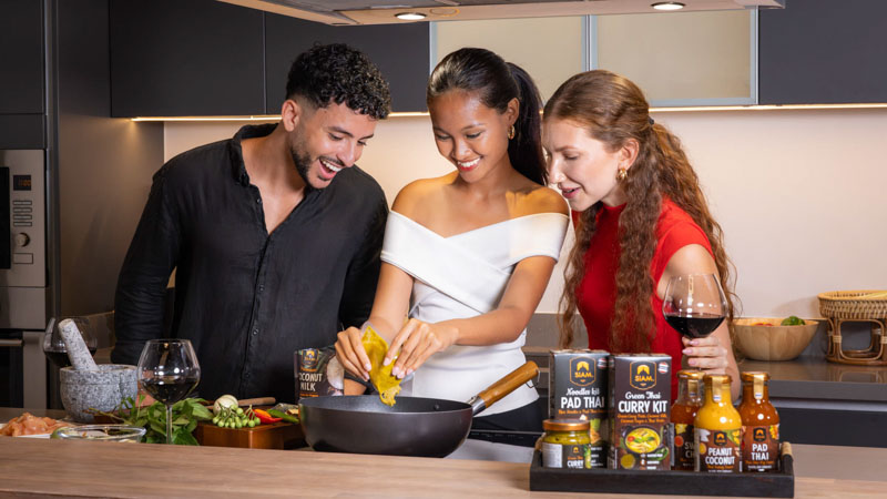Trois personnes souriantes préparent un repas ensemble dans une cuisine moderne, avec des ingrédients et des kits de cuisine sur le comptoir.