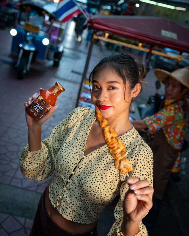 Jeune femme souriante tenant une bouteille de sauce chili sucrée dans une main et une brochette de poulet dans l'autre, dans une rue animée avec un tuk-tuk en arrière-plan.