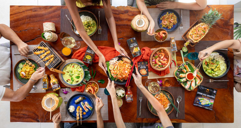 Vue plongeante d'un groupe de personnes partageant un repas avec des plats thaïlandais colorés et des boissons sur une grande table en bois.
