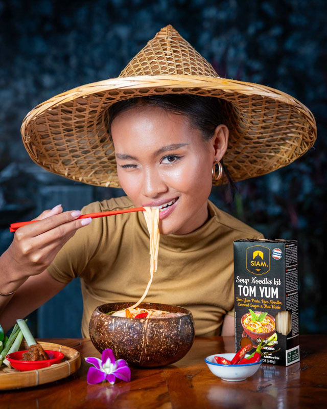 Femme asiatique portant un chapeau en bambou mangeant des nouilles Tom Yum avec des baguettes devant un kit de soupe Tom Yum Siam sur une table en bois.