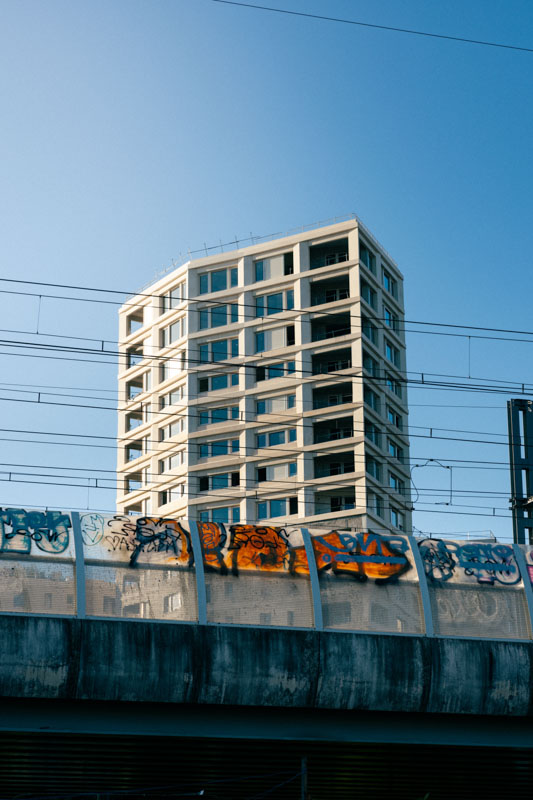 Immeuble moderne en béton vu derrière un pont orné de graffitis colorés sous un ciel bleu clair.