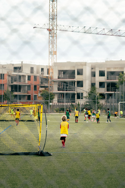 Des enfants jouent au football sur un terrain extérieur avec des bâtiments en construction et une grue en arrière-plan.