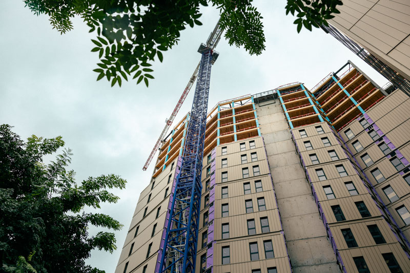 Grue de construction bleue devant un immeuble en construction avec des feuilles d'arbres au premier plan sous un ciel nuageux.