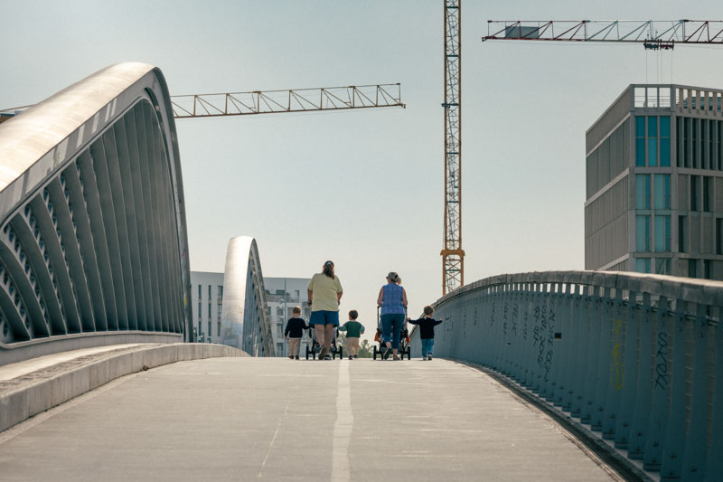 Des adultes et des enfants marchant sur un pont moderne avec des grues de construction en arrière-plan.