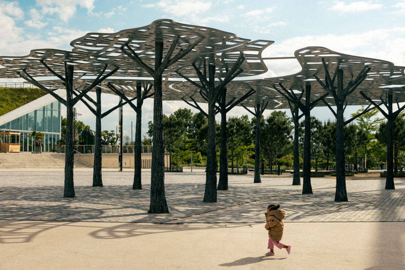 Enfant marchant près d'une structure métallique moderne en forme d'arbres dans un espace urbain avec des arbres verts en arrière-plan.