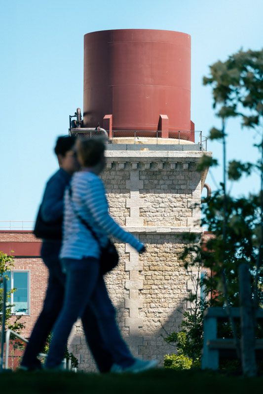 Deux personnes marchent devant une grande citerne cylindrique en pierre avec une partie supérieure rouge sur un ciel bleu.