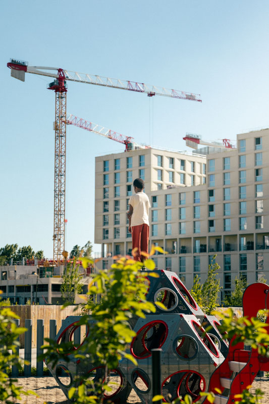 Un enfant debout sur une structure de jeu géométrique dans une aire de jeux avec des bâtiments modernes et des grues en arrière-plan sous un ciel bleu clair.